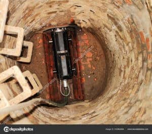 A submerged sump pump inside a deep, cylindrical, brick-lined well with visible iron ladder rungs on the side.