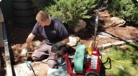 A man repairing outdoor plumbing with tools and equipment on a sunny day in a landscaped garden area.