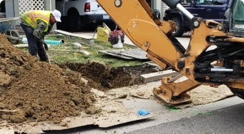 Construction worker supervising an excavator digging up a city street, with piles of dirt and construction materials around.