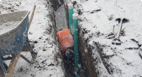 Underground pipeline installation in a snow-covered trench with a blue pipe, red valve, tools, and a wheelbarrow nearby.