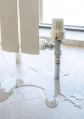 A radiator leaking water onto a wooden floor beside a window, with bright daylight streaming in.