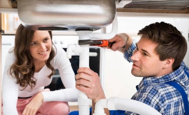 A man and a woman fixing a sink's plumbing, with the man using a wrench while the woman watches.