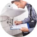 A man inspecting the plumbing under a sink while making notes on a clipboard.