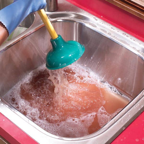 A person wearing a blue glove uses a green plunger in a stainless steel sink filled with soapy water.