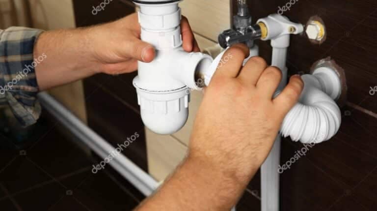 A plumber's hands installing a white p-trap under a sink, connecting it to the sink's drainage pipes.