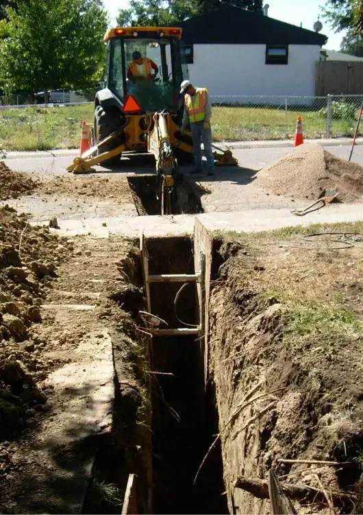 Construction workers using a backhoe to dig a trench on a residential street, with cones marking the work area and a ladder placed inside the trench.