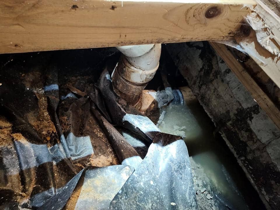 A leaking pipe under wooden beams with accumulated water and debris around it in a crawl space.