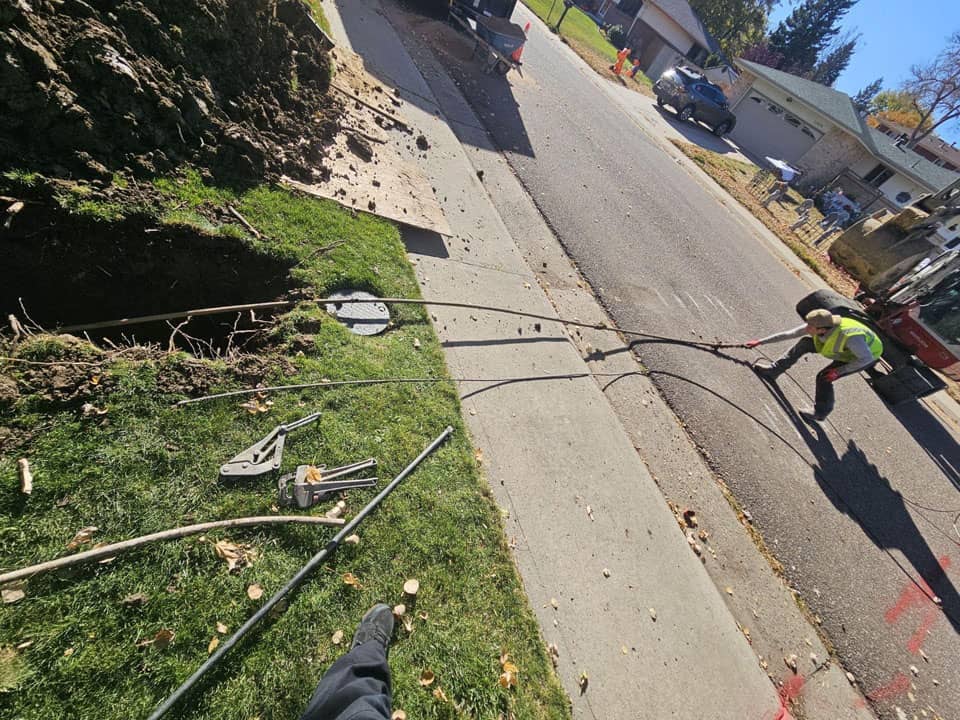 A worker in a fluorescent vest pulls cables from a trench across a residential street. Tools and equipment are scattered on the grass beside the sidewalk.